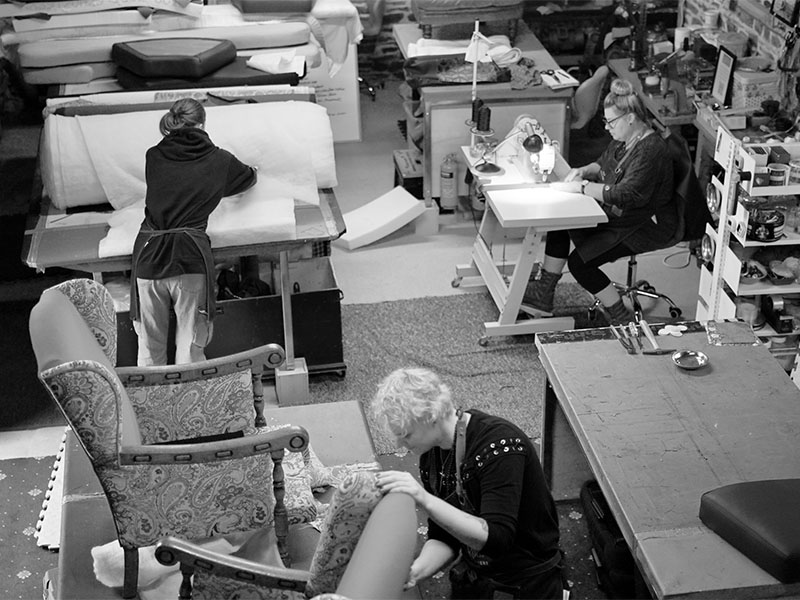 About Us: The Needle Rock team working on upholstery projects in the workshop, with sewing and crafting stations in a busy creative space.